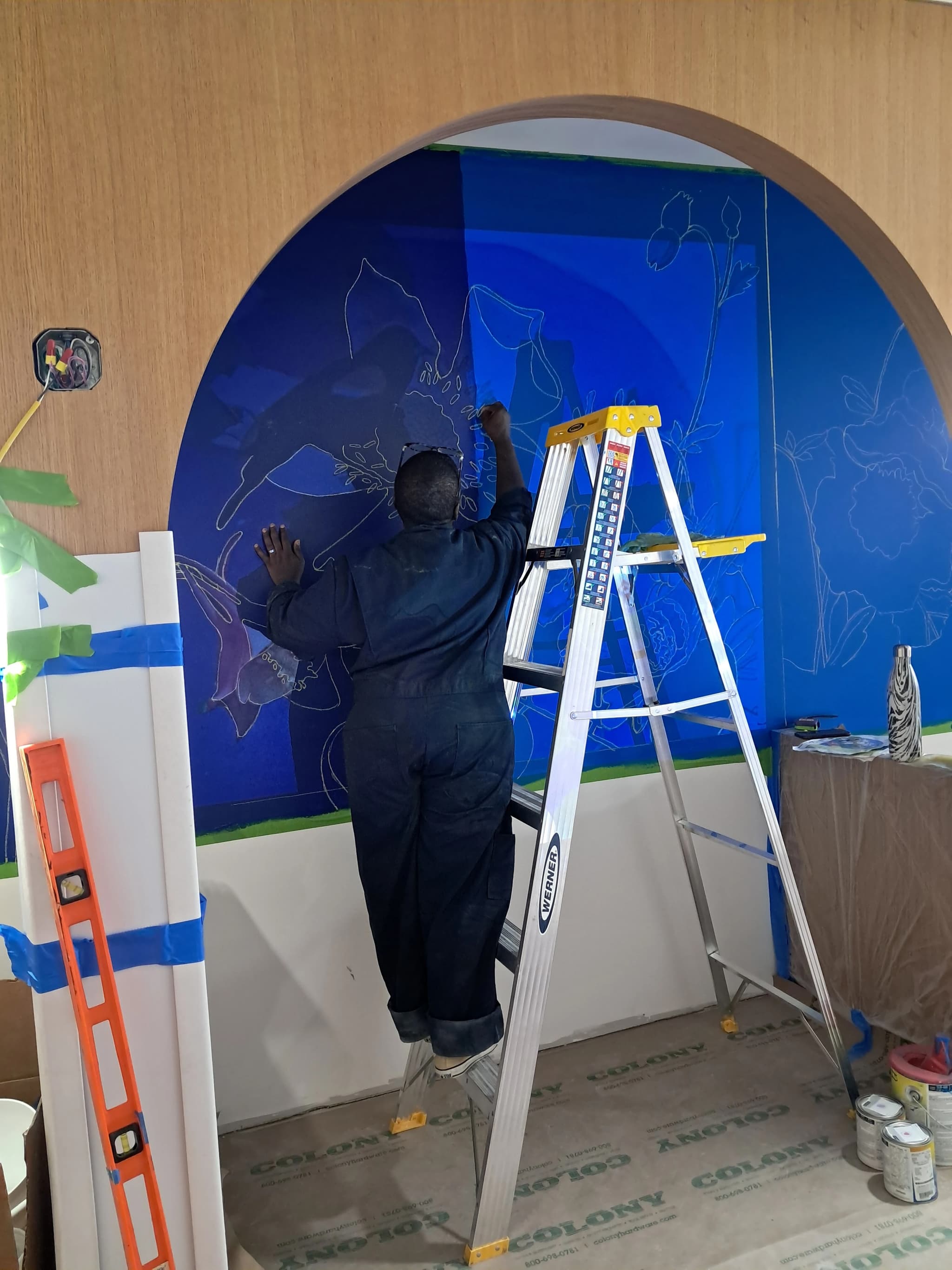 A muralist in dark coveralls stands on an aluminum step ladder, sketching floral outlines onto a bright blue painted wall within an arched wooden alcove, with painter's tape, drop cloths, and supplies scattered around the workspace