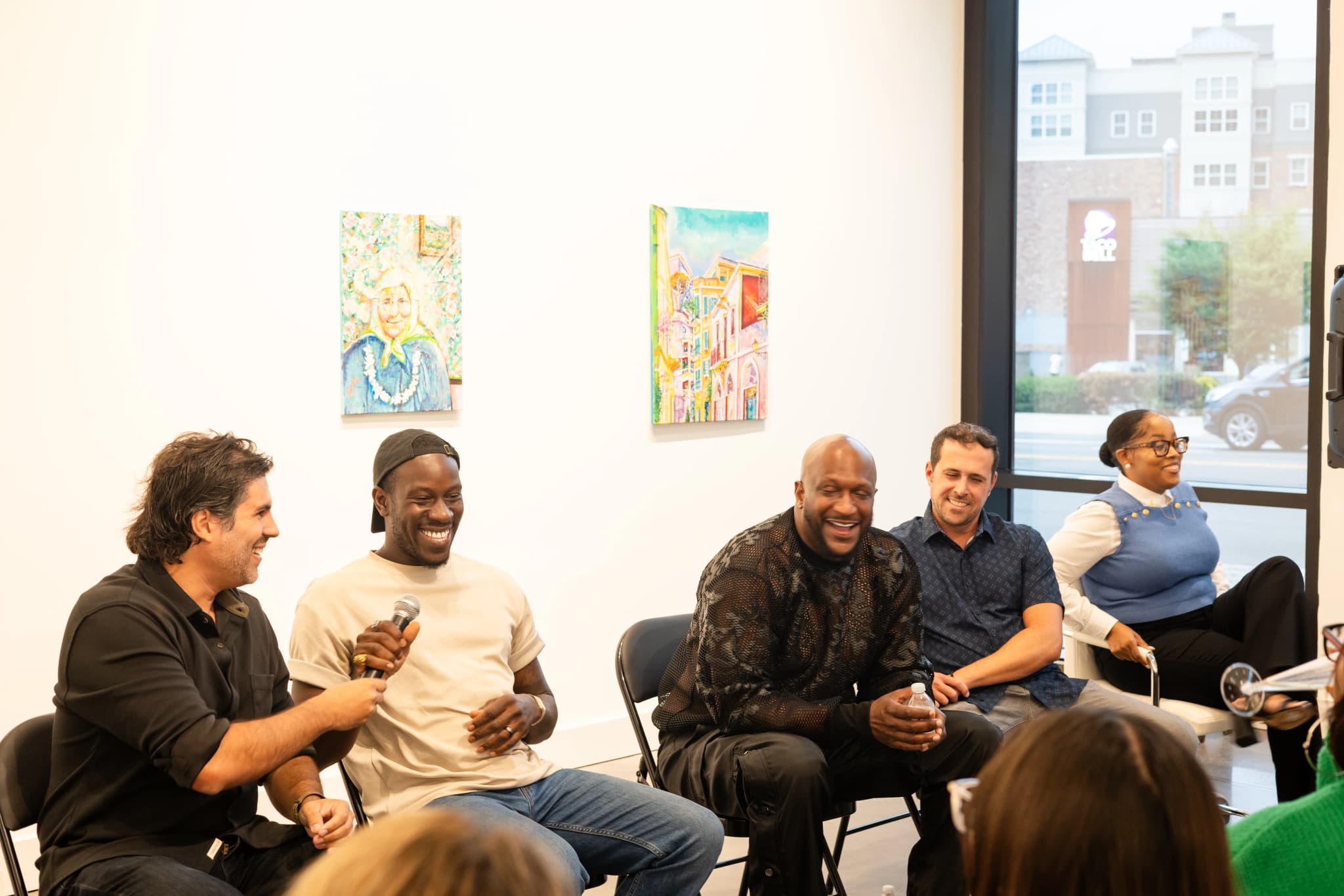 Four panelists seated in folding chairs at a gallery laugh together during a film panel discussion, with one man holding a microphone, two colorful paintings on the white wall behind them