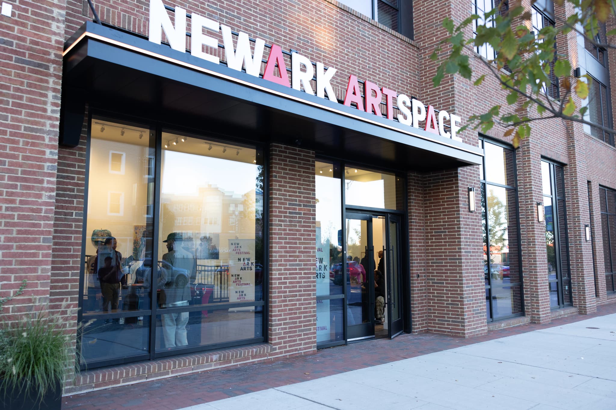 Exterior of the Newark ArtSpace gallery on a brick building, with large "NEWARK ARTSPACE" signage in white and pink above the entrance. Newark Arts Festival posters are visible through the front windows, and people can be seen gathered inside.