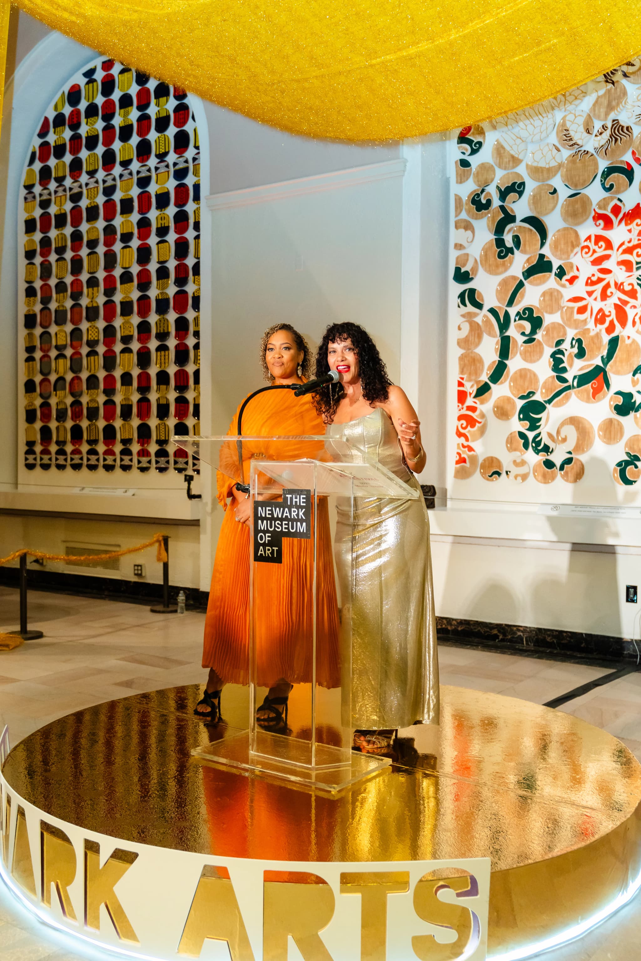 Two women stand at a clear acrylic podium marked "The Newark Museum of Art," one in a flowing orange dress and one in a shimmering gold gown speaking into the microphone, on a circular gold stage bordered with "NEWARK ARTS" lettering, with a glittering yellow canopy overhead and colorful patterned artworks framing the wall behind them