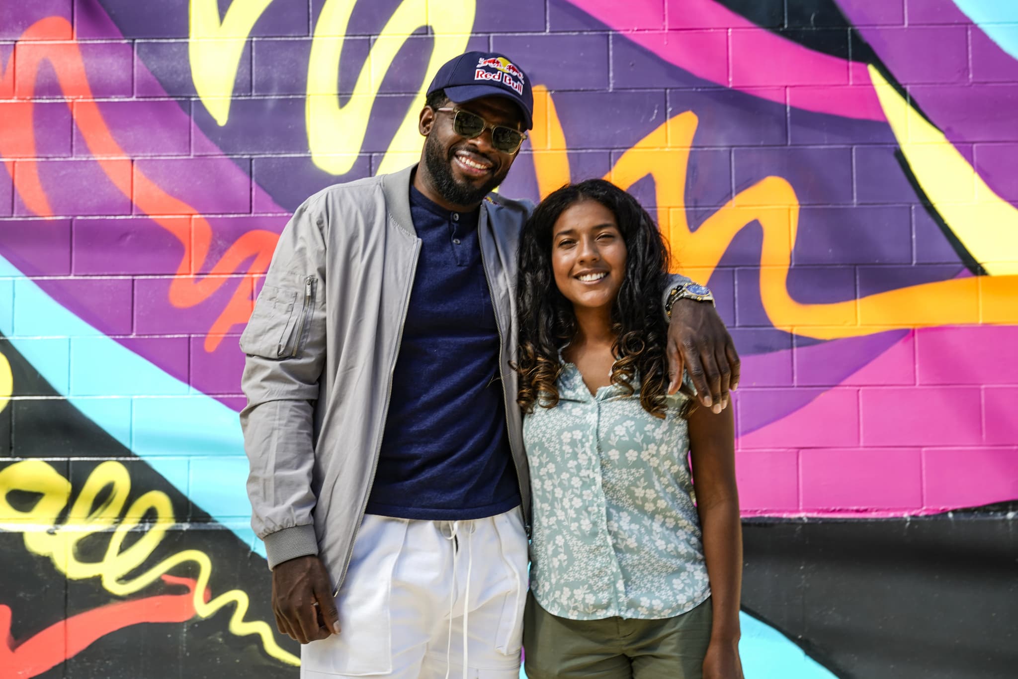 A tall man in a Red Bull cap, sunglasses, gray bomber jacket, and white pants stands with his arm around a smiling young woman with long curly hair in a light floral blouse, both posing in front of a vibrant graffiti mural in purple, pink, yellow, and blue at the P.K. Subban mural unveiling.